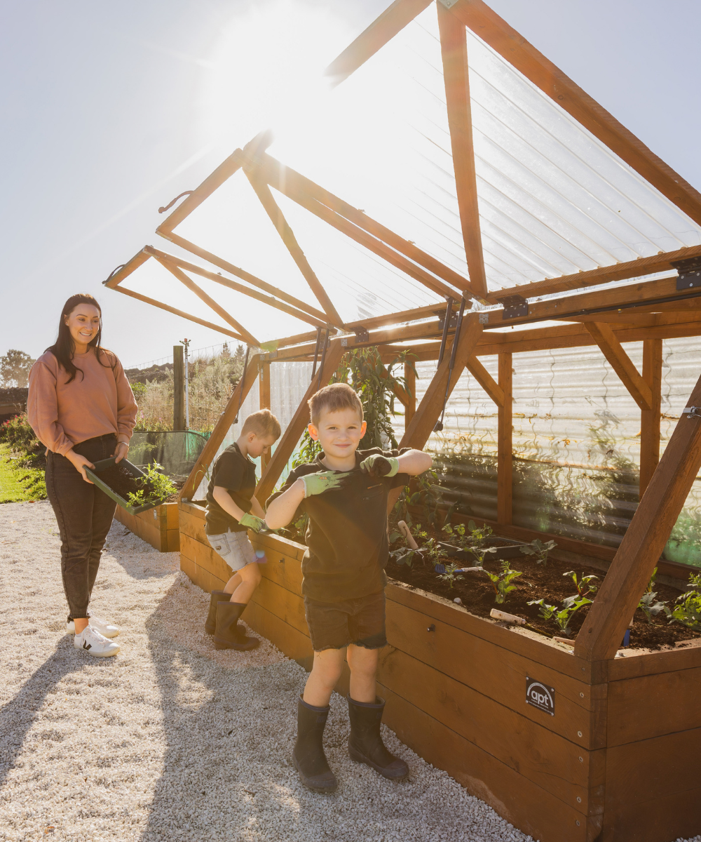 A family gardening a raised garden bed with a translucent roof under bright sunlight. A woman stands on the left holding a hose, a young boy stands in the foreground holding a garden tool, and another child sits on the raised garden bed.